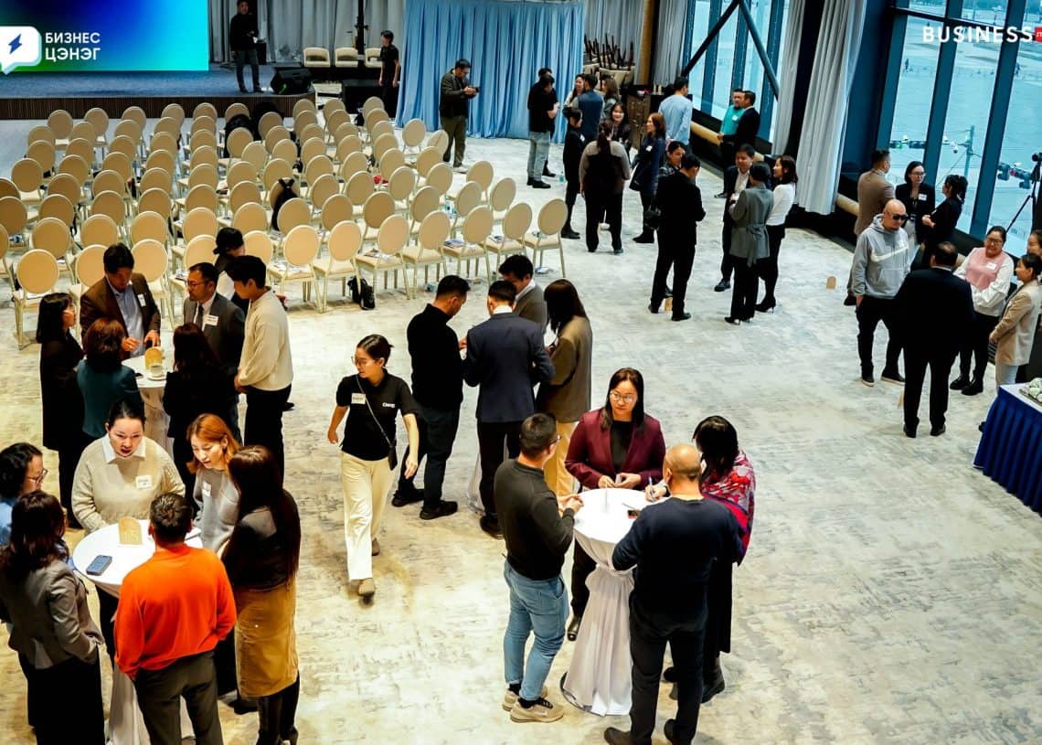 A well-lit conference room with people mingling around cocktail tables. Rows of empty chairs are set up in the background. A large screen displays content, and business professionals are networking and conversing in small groups.