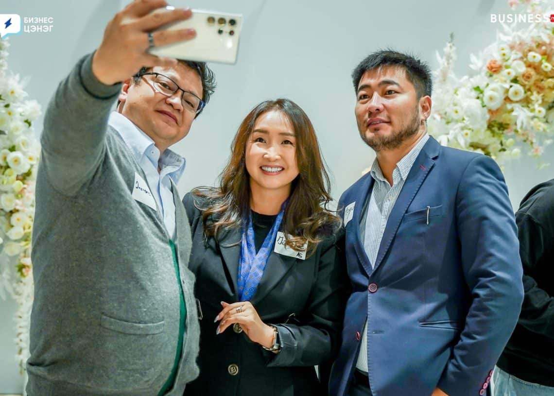 Three people are taking a selfie together. The group includes two men and one woman, all smiling. They are indoors, and the background features floral decorations.