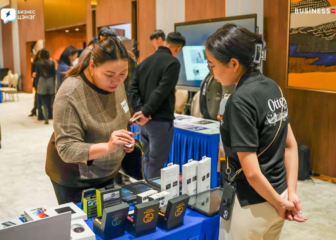 Two people stand at a table displaying electronic gadgets. One person is demonstrating a product to the other. The background shows more people and display tables in a conference setting.