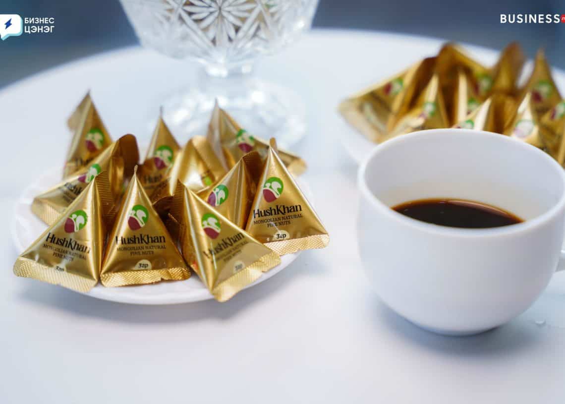 A white cup filled with coffee is placed next to a plate of individually wrapped gold pyramid-shaped butter packets labeled "HashKhan." A decorative dish is partially visible in the background.