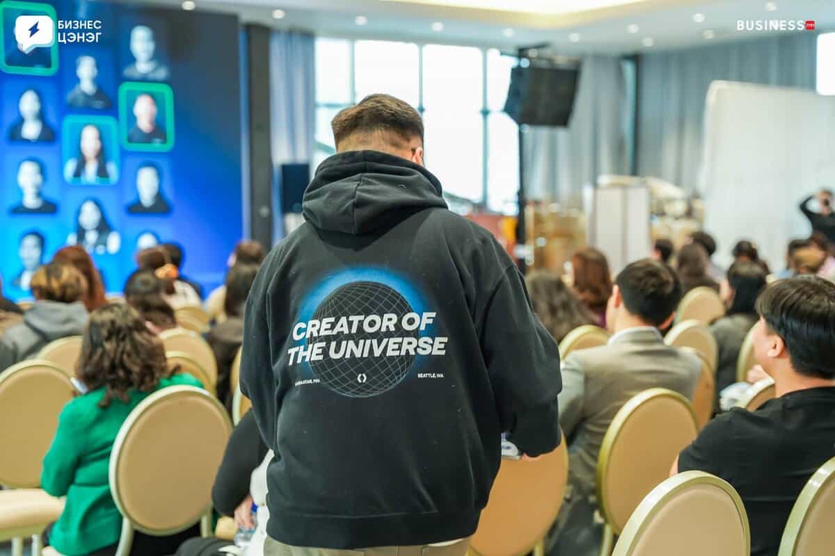 A person wearing a black hoodie with "CREATOR OF THE UNIVERSE" printed on the back stands in an auditorium. The room is filled with seated attendees facing a stage displaying a large screen with graphics and photos.