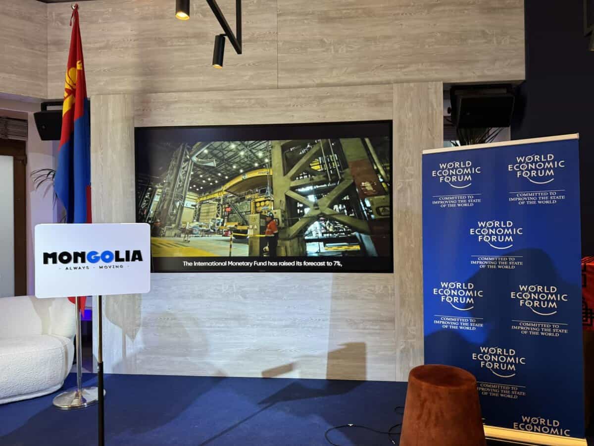A stage with a Mongolian flag on the left, a screen displaying an industrial scene, and a banner with "World Economic Forum" logos on the right. A sign with "Mongolia" and a slogan is in front of a white chair.
