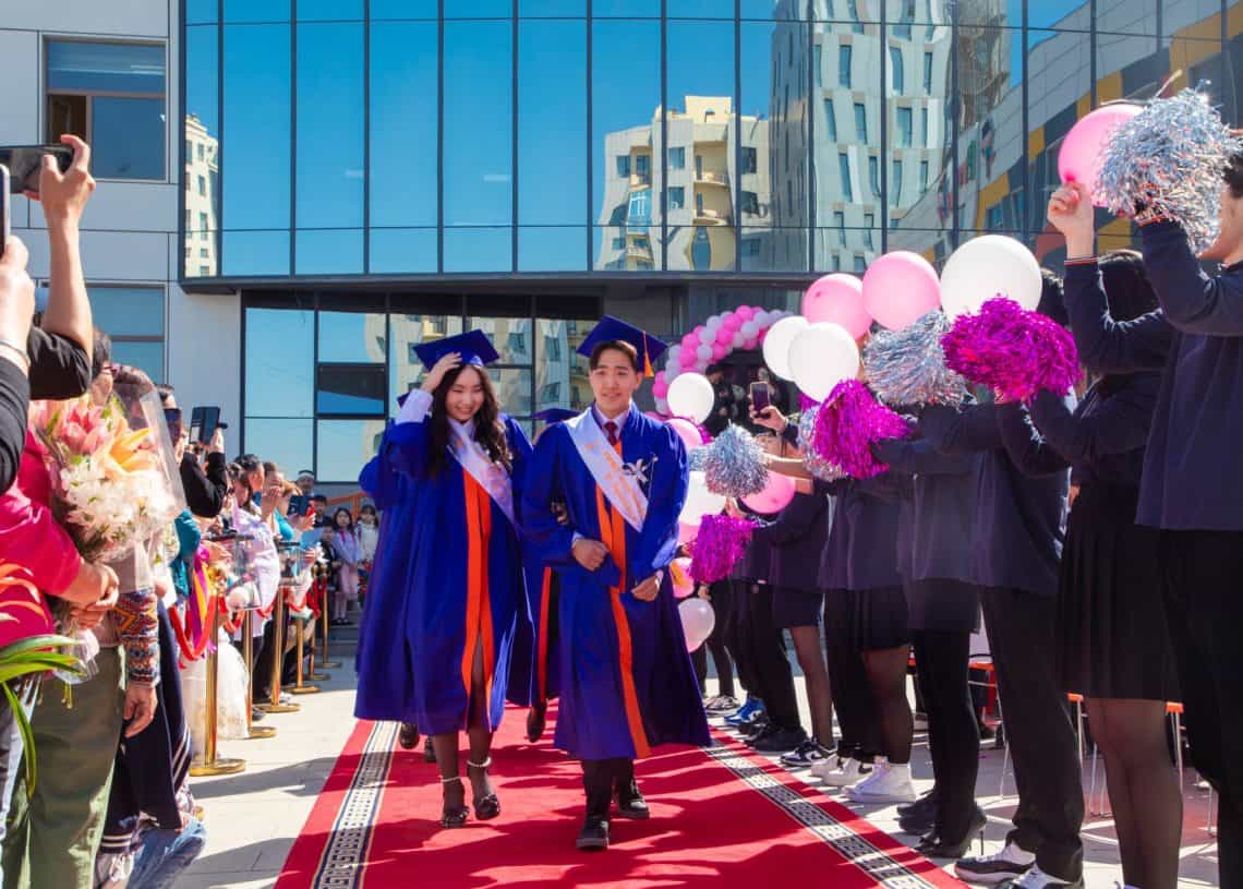 Two graduates in blue gowns walk down a red carpet lined with cheering people holding pom-poms. They smile as they approach a glass building adorned with balloons. A festive atmosphere surrounds the graduation ceremony.