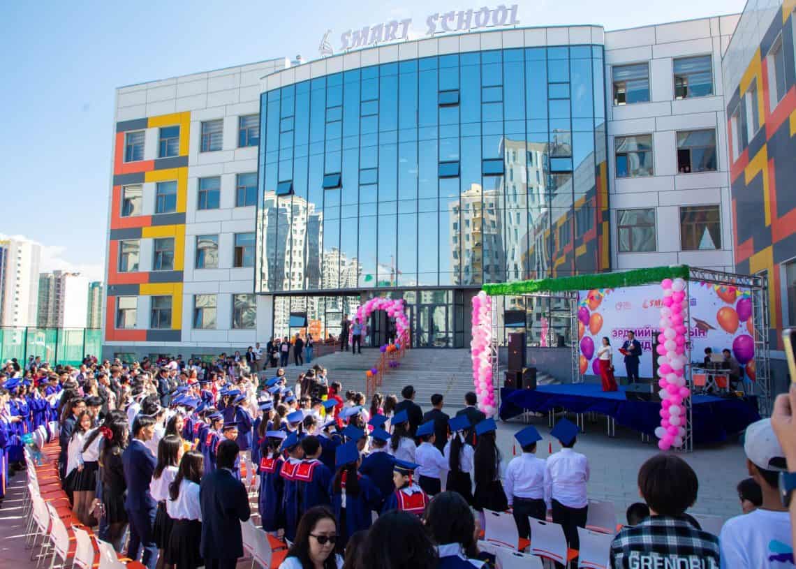 A graduation ceremony outside a modern school building with a glass facade. Graduates in blue gowns and caps sit while an audience watches. A stage is decorated with pink balloons, and a speaker addresses the crowd.