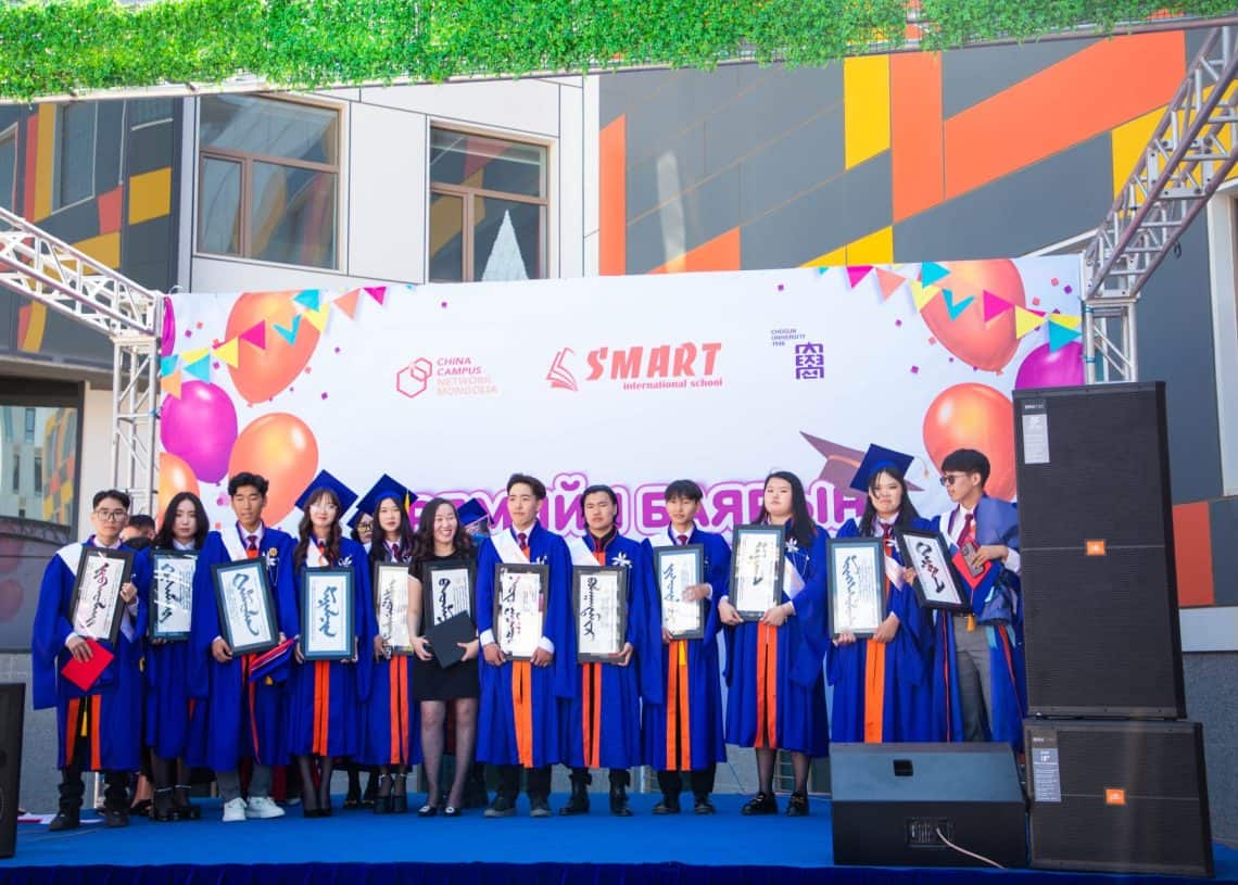 A group of graduates in blue and orange robes stand on a stage holding certificates. They are in front of a colorful backdrop with balloons and logos. The event is outdoors with a building in the background.