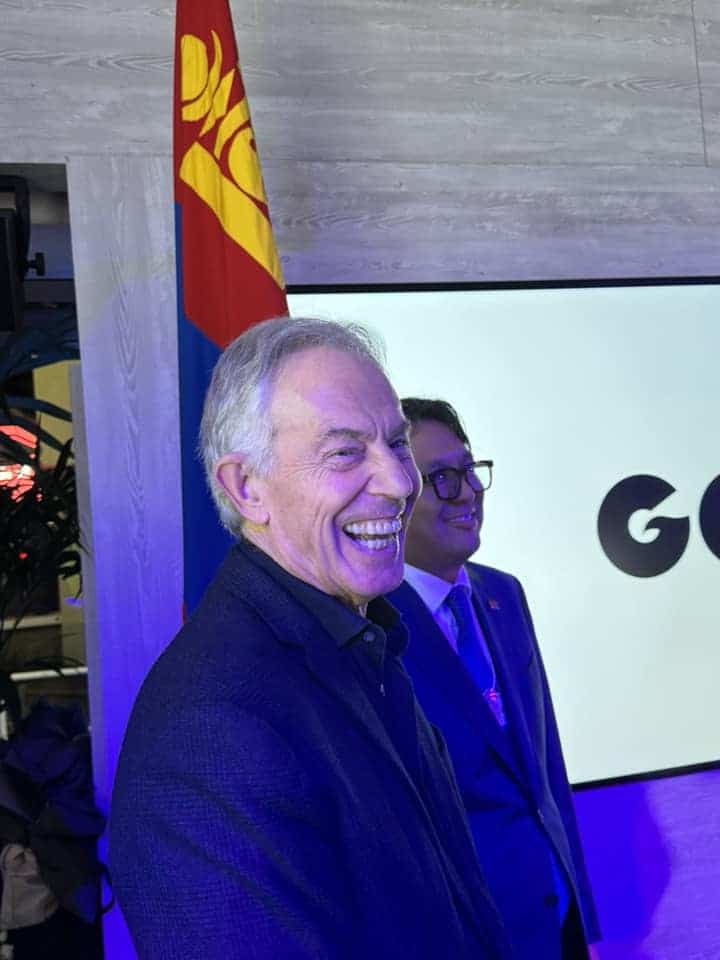 Two people stand in a room with a Mongolian flag. One is smiling broadly, and the other is in the background, facing forward. They are lit with blue lighting, and there is a screen behind them.
