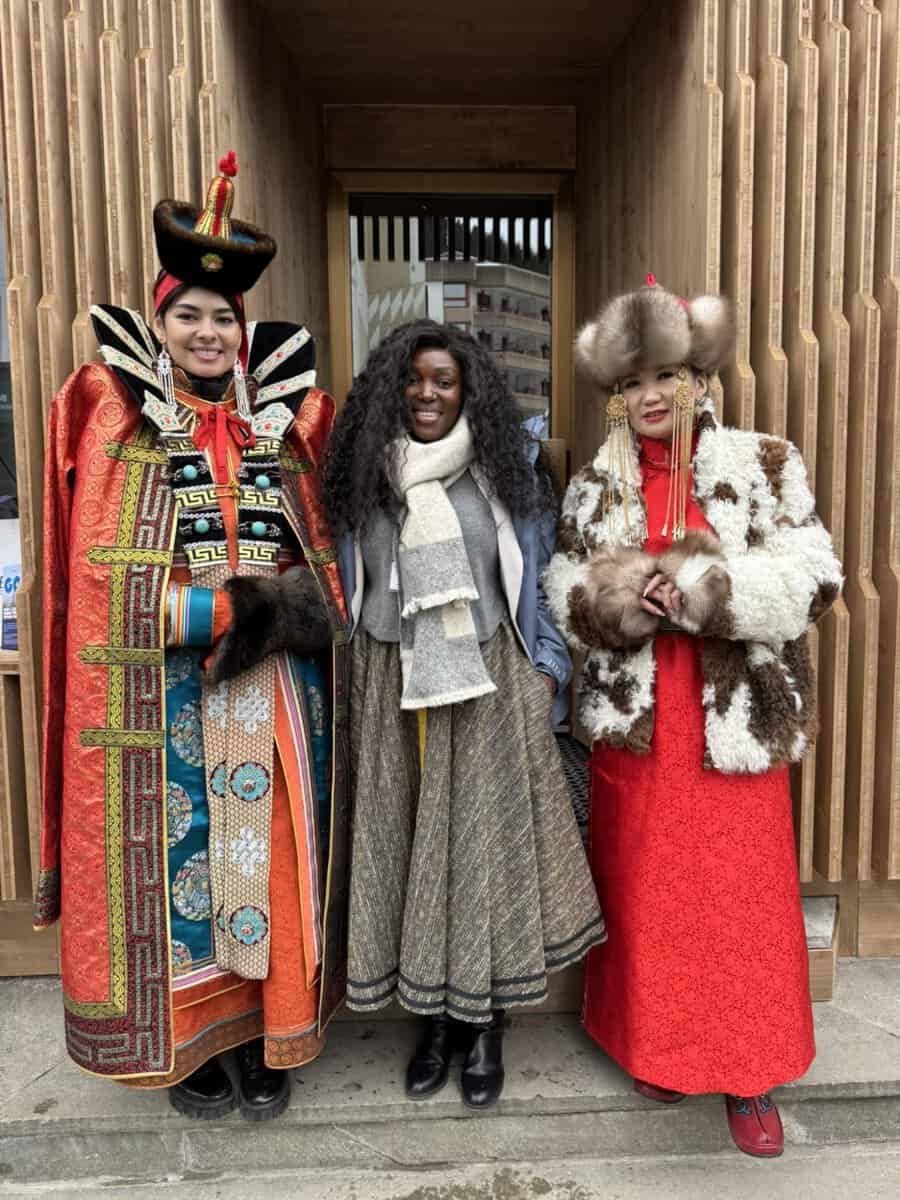 Three people stand in front of a wooden building. Two wear traditional, colorful Mongolian attire with ornate hats, and one in the center wears a grey scarf and coat. They are smiling and standing close together.