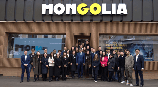A diverse group of people stands in front of a building with a sign that reads "MONGOLIA" in bold letters. They are smiling and posing for the picture on a paved area.