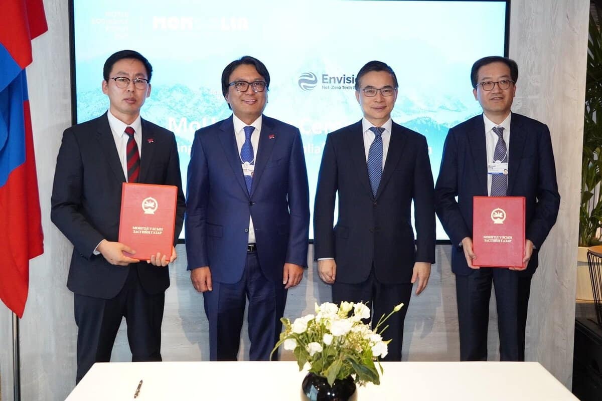 Four men in suits stand together in front of a display screen reading "Envision" and "Mongolia." Two men on the left hold red folders. On a table in front of them is a vase with white flowers. Flags are partially visible on the sides.