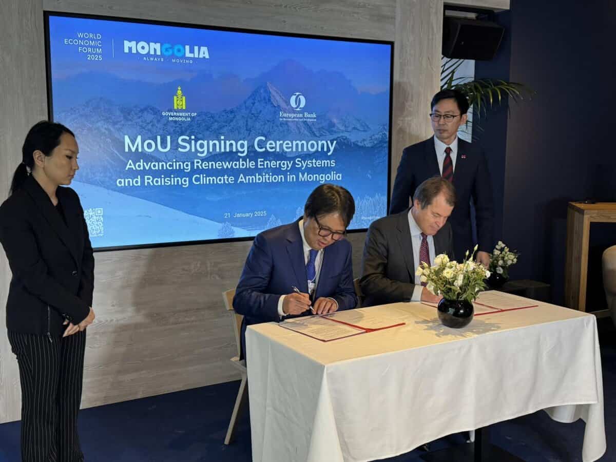 Four individuals are at a table during an MoU signing ceremony. Two are seated and signing documents, while two stand observing. A sign in the background mentions "Advancing Renewable Energy Systems and Raising Climate Ambition in Mongolia.