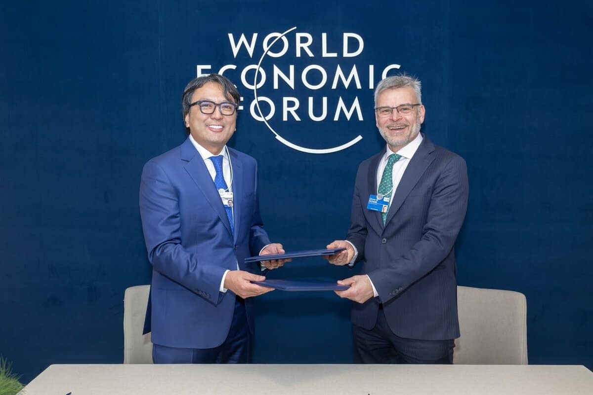 Two men in suits shake hands while holding a document at the World Economic Forum. They are standing in front of a large logo on a blue background, smiling towards the camera.