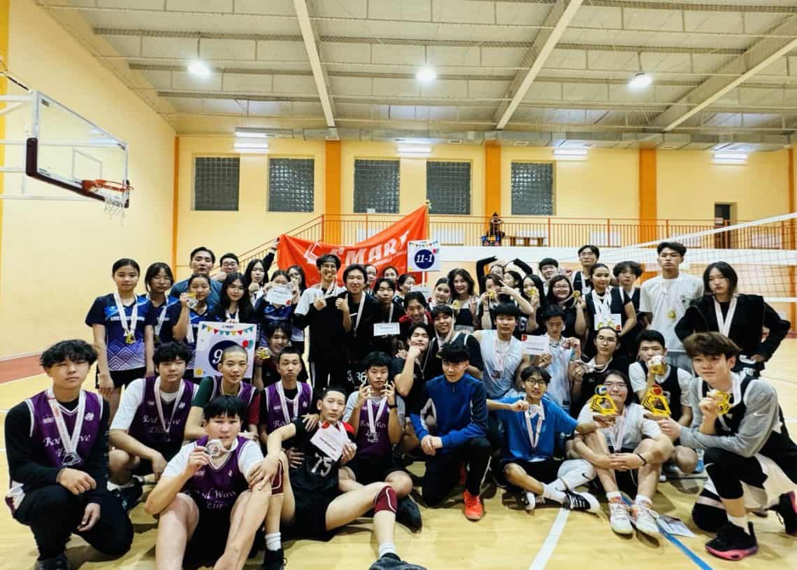 A large group of young people, some wearing sports uniforms and holding medals, pose for a group photo in an indoor sports hall. A basketball hoop is visible, and a bright orange banner is in the background.