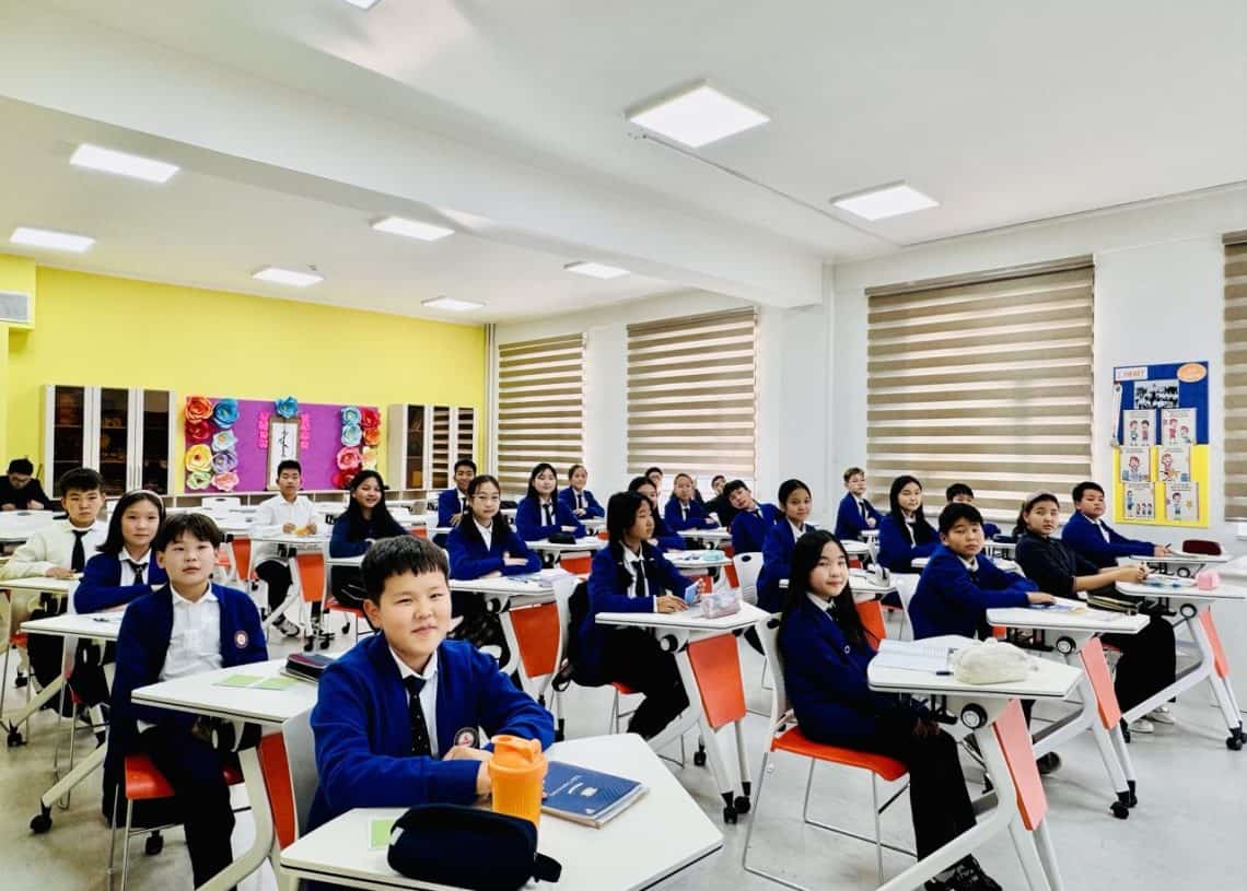 A classroom filled with students sitting at desks, wearing blue sweaters and white shirts. The classroom has bright walls with decorations and a whiteboard. The students are facing forward, with smiles and attentive expressions.