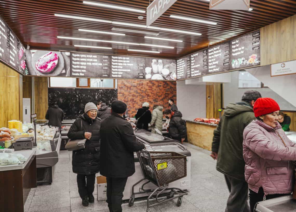 People shopping in Дүүхээ дэлгүүр, a market with a wooden ceiling and fluorescent lights illuminating various food displays. Some wear winter clothing like coats and hats. Signs with food images hang from above, and a shopping cart waits in the foreground.