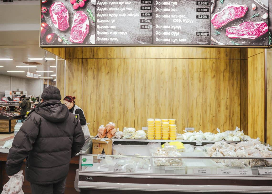 A person in a winter coat stands at the meat counter of Дүүхээ дэлгүүр. Behind the counter, a worker arranges packaged products and yellow containers. Above, a digital menu lists items and prices in Russian, all under the store's bright lighting.