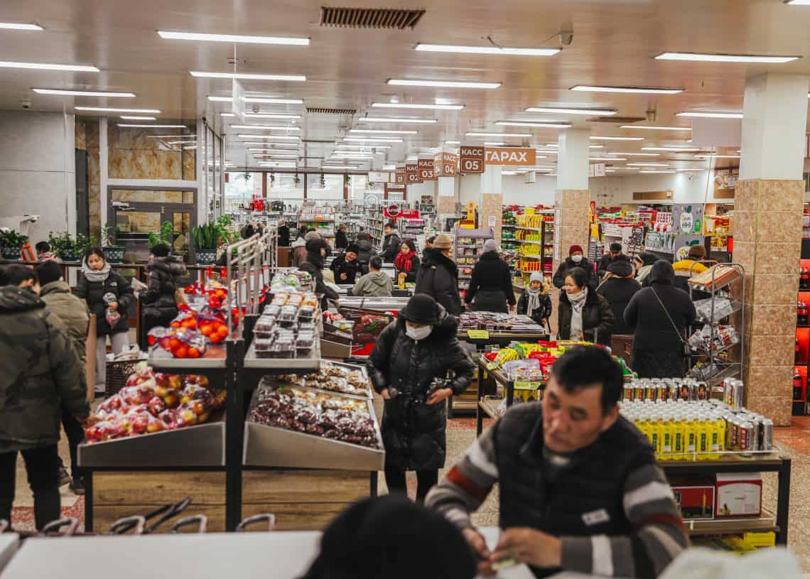 In a bustling Дүүхээ дэлгүүр, shoppers in winter clothing browse aisles brimming with products. As some pick items, others wait at the checkout. Bright lighting and organized shelves enhance the lively atmosphere of this vibrant supermarket scene.