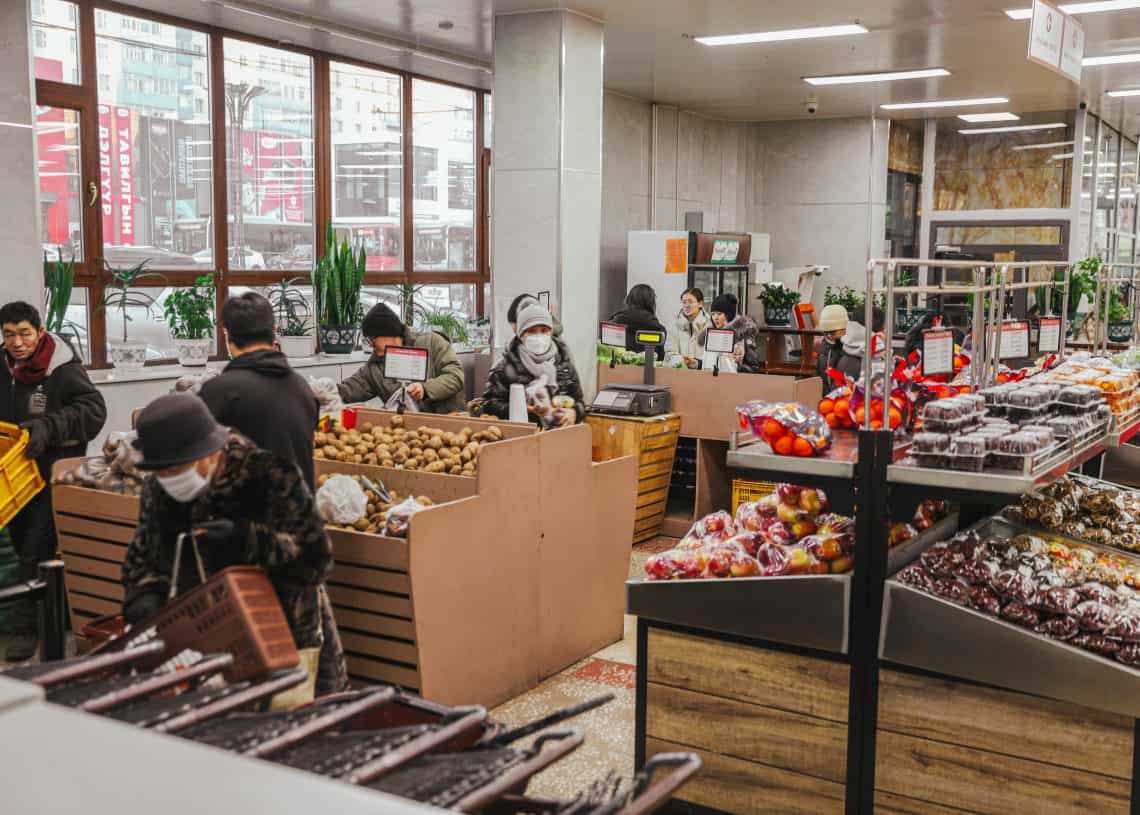 Shoppers browse a well-lit Дүүхээ дэлгүүр, where fresh fruits and vegetables fill the bins. Some wear masks as they carefully select their produce, with sunlight streaming through large windows casting a warm glow on the bustling scene.