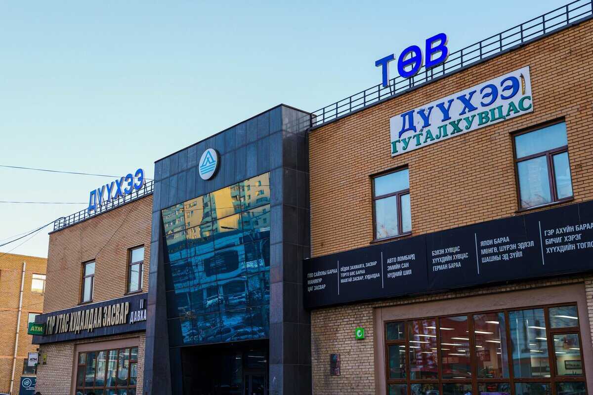 A brick building with a digital screen displaying a cityscape. Blue signs with Mongolian script are on the roof and walls. There's a glass doorway at the entrance. The building appears to be a commercial or public center.