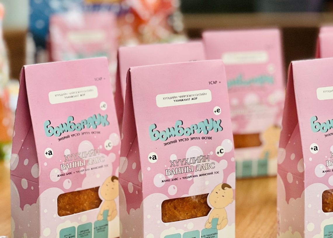 Several pink and white packaged candy boxes are displayed on a wooden table. The design features a cartoon baby. A blurred background shows more packaged items and plants.