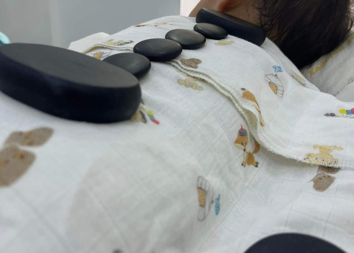 A person lying down with several black hot stones placed on their back, covered by a white blanket with cartoon designs. The setting appears to be a spa or relaxation environment.