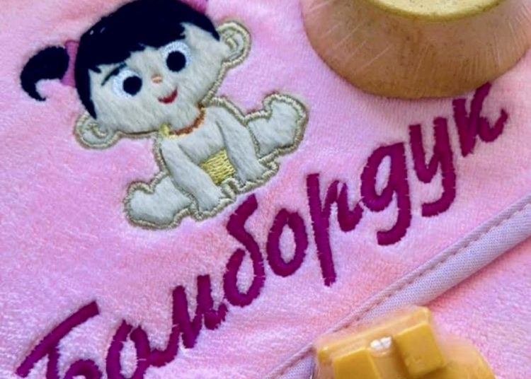 Pink towel featuring an embroidered cartoon baby with pigtails and the text "Бомбардир." Nearby, a round soap with a lavender label and a yellow soap shaped like a vehicle are visible.