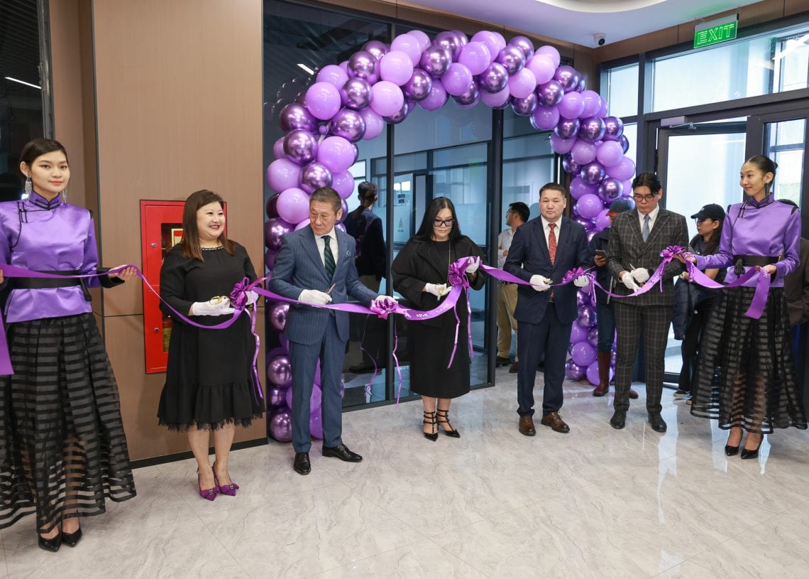 A group of people in formal attire cut a purple ribbon at an opening ceremony, standing under a purple balloon arch indoors. Two women in purple outfits stand at each end of the ribbon.