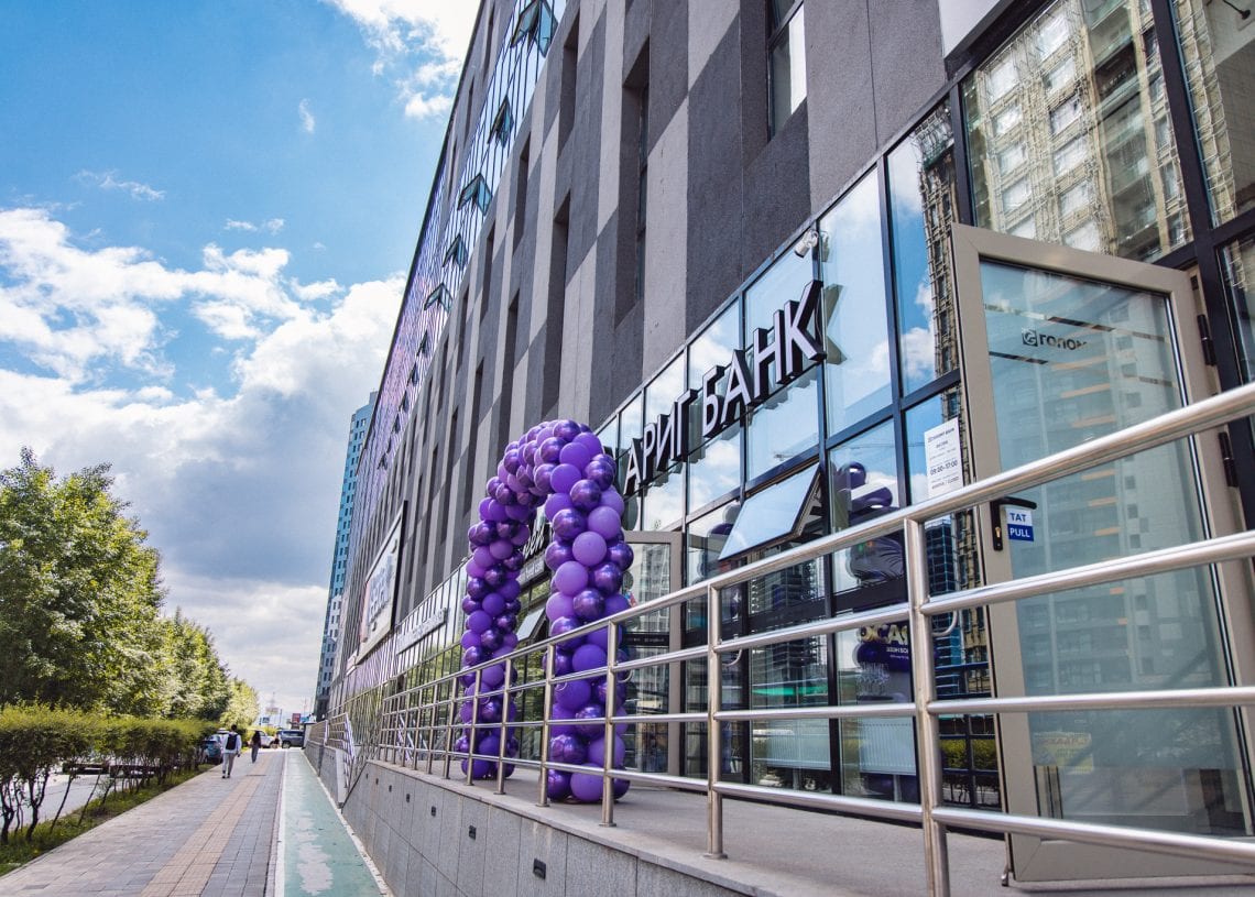 A modern bank building with large windows and a glass door. A decorative arch of purple balloons stands at the entrance, and a metal ramp leads up to the door. Trees and a sidewalk run alongside the building.