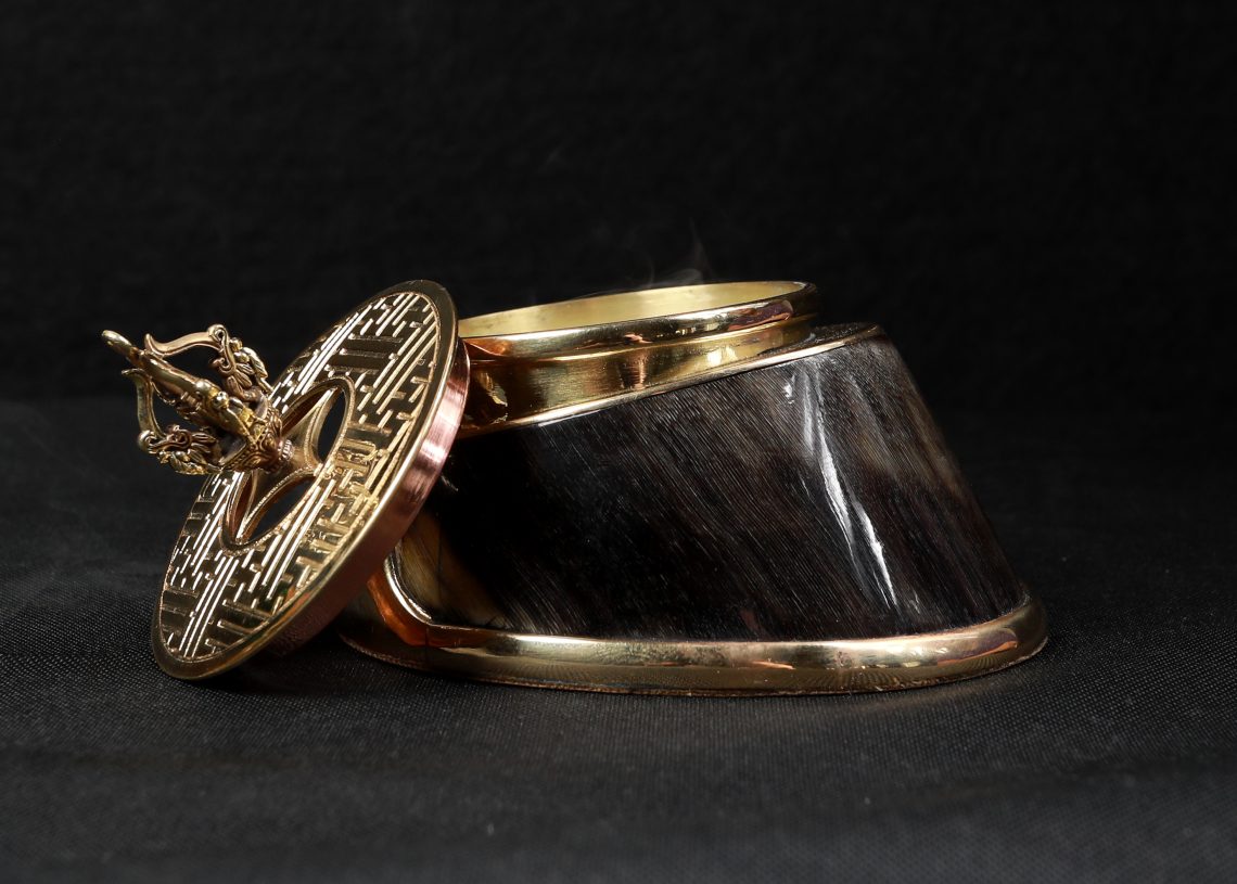 Decorative brass and horn box with lid partially open on a dark background. The lid features intricate geometric patterns and a small handle. The main body of the box has a glossy, dark horn texture with brass trim.