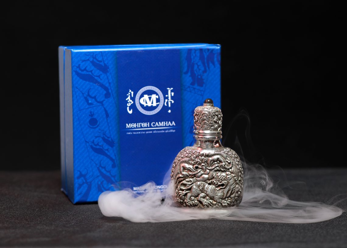 An intricately detailed silver bottle sits in front of a blue box with white text and symbols. The bottle is adorned with dragon engravings and is surrounded by wisps of fog, set against a dark background.