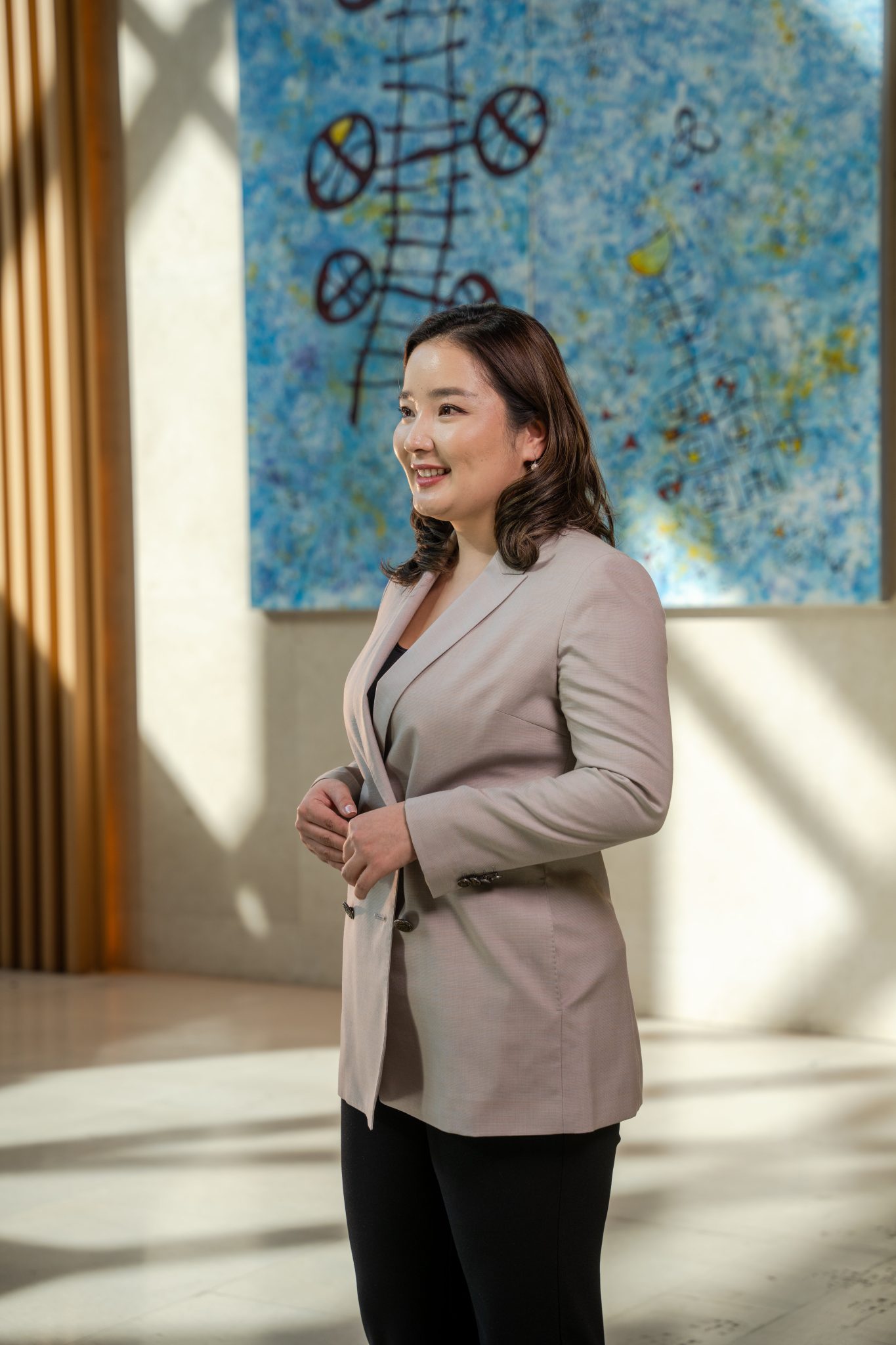 A person in a beige blazer stands in a sunlit room, smiling, with hands clasped. Behind them, a colorful abstract painting featuring circular and linear elements reflects the innovative spirit of Дижитал Эдү Менежмент.