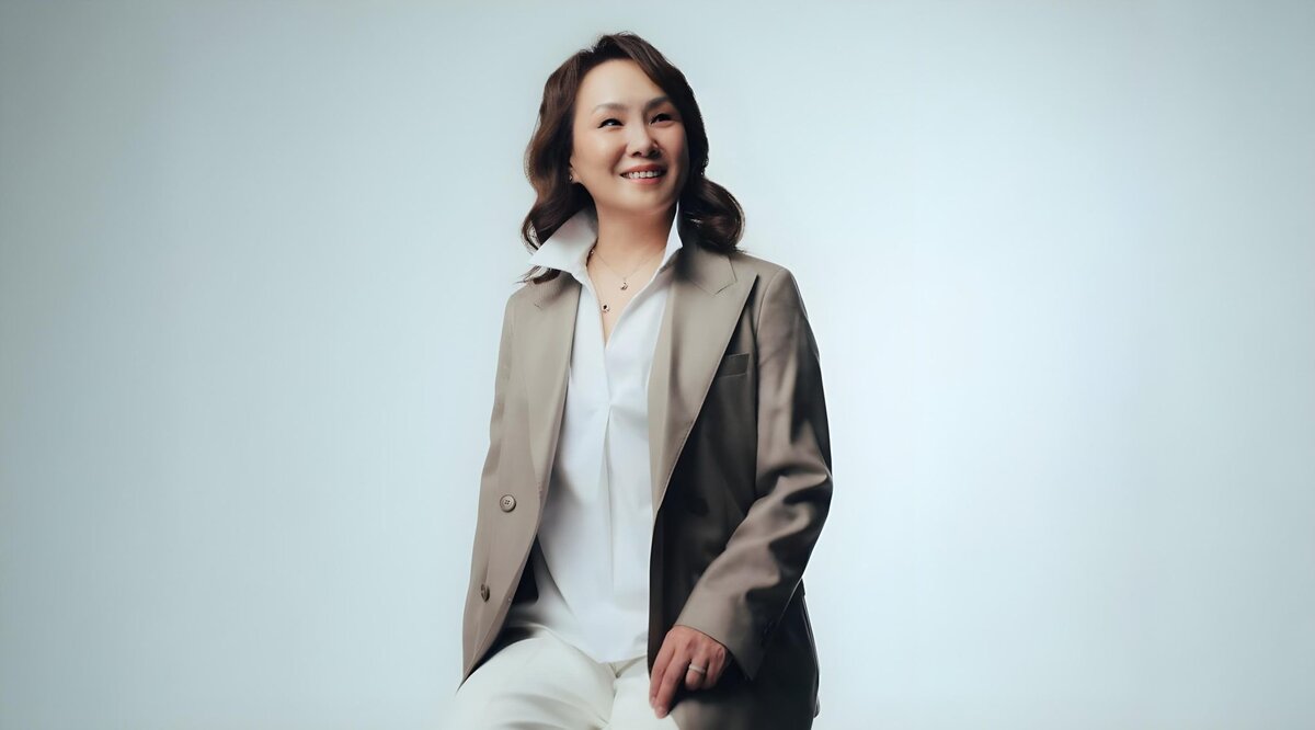 A person with shoulder-length hair sits on a stool against a light gray background, wearing a beige blazer over a white blouse and pants, smiling slightly to the side.