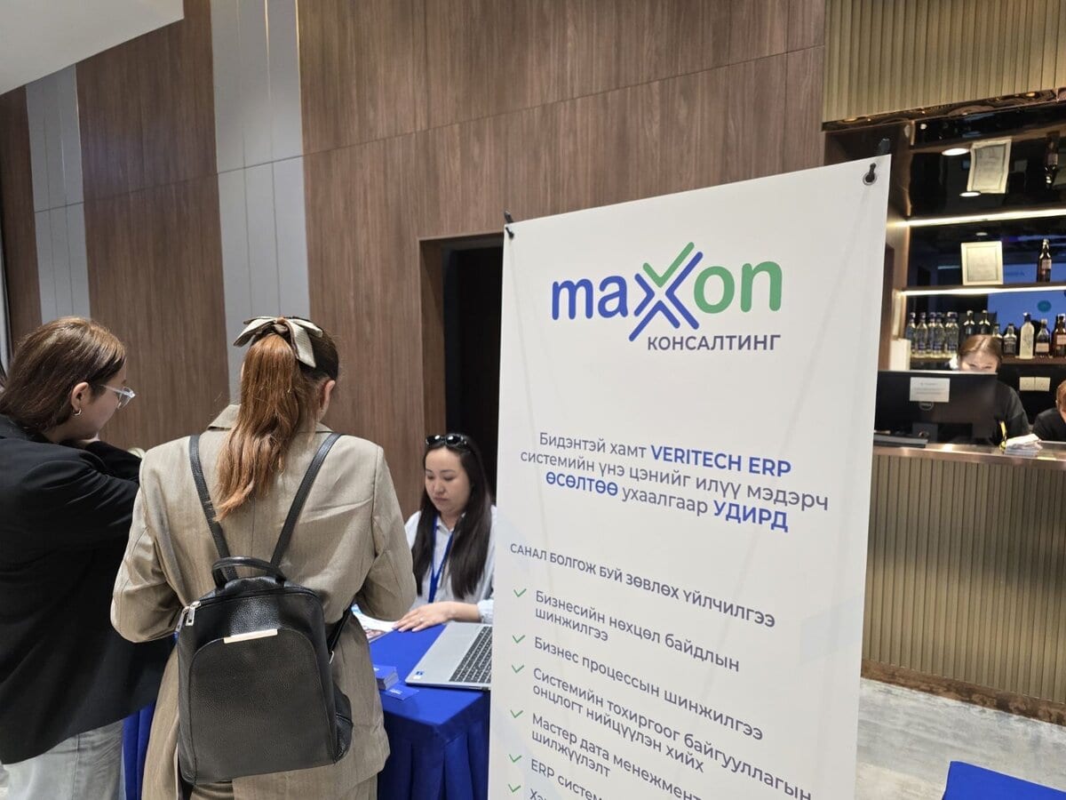 Two women stand at a registration desk near a banner for Maxxon Consulting and VERTEREN ERP in Mongolian at an indoor event. A staff member is seated with a laptop behind the desk. In the background is a bar area.