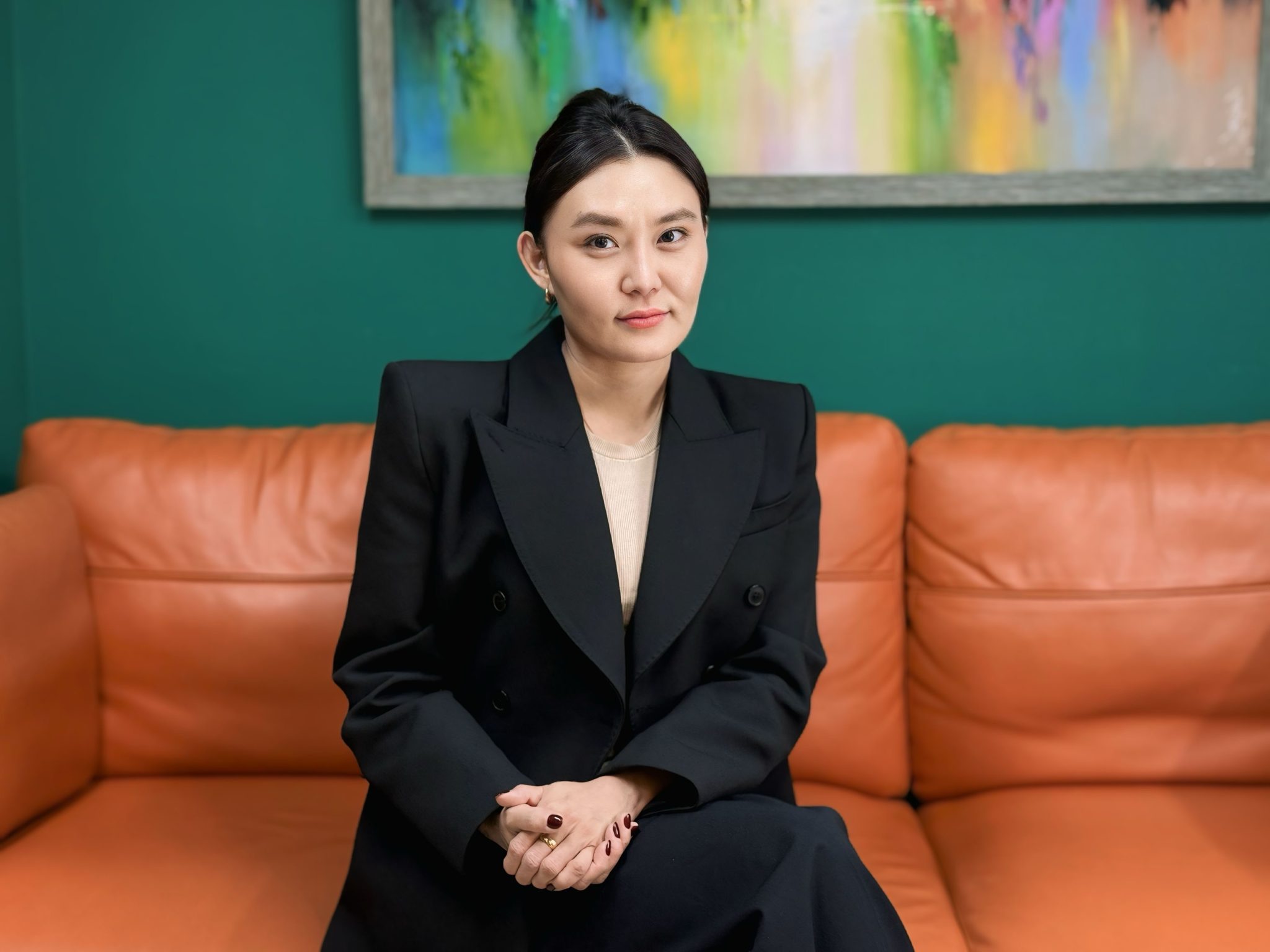 A woman in a black blazer sits on an orange couch against a green wall with a colorful abstract painting behind her, looking at the camera with a slight smile.