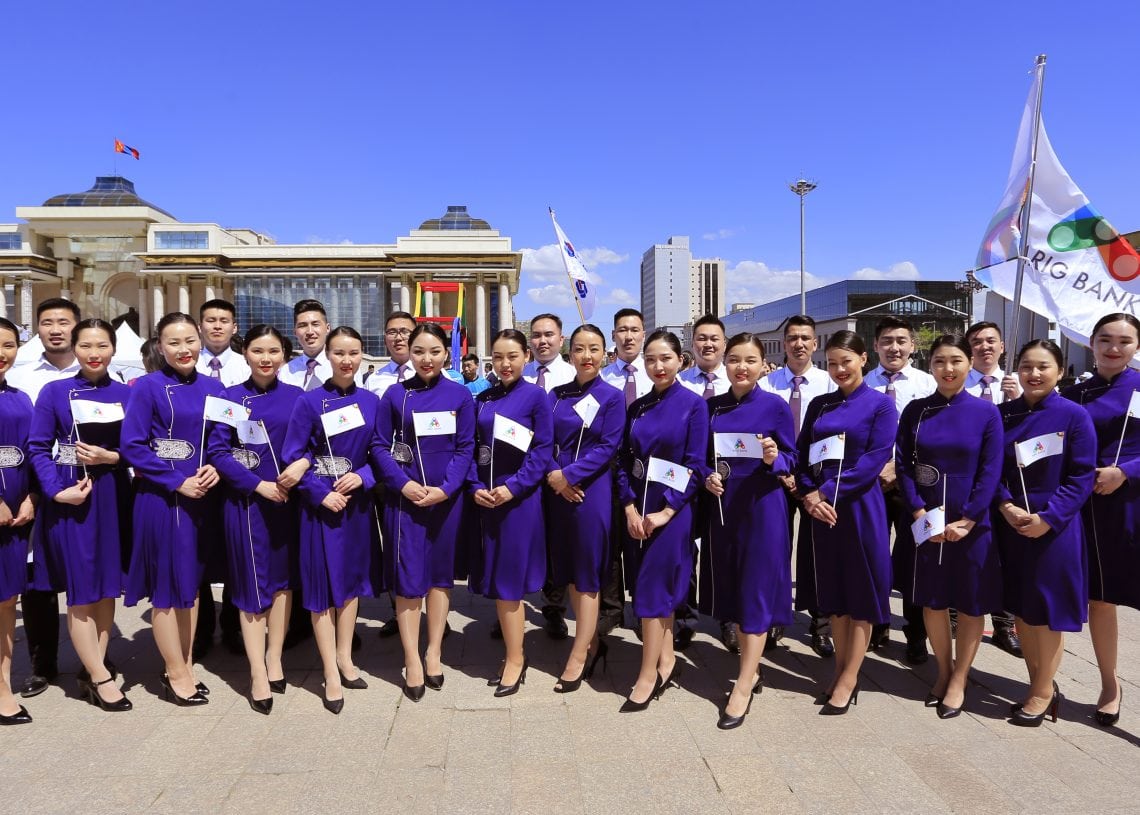 A group of women in matching purple dresses stand in a line outdoors, holding small white flags. Behind them are large buildings, blue sky, and a person holding a white flag with a logo.
