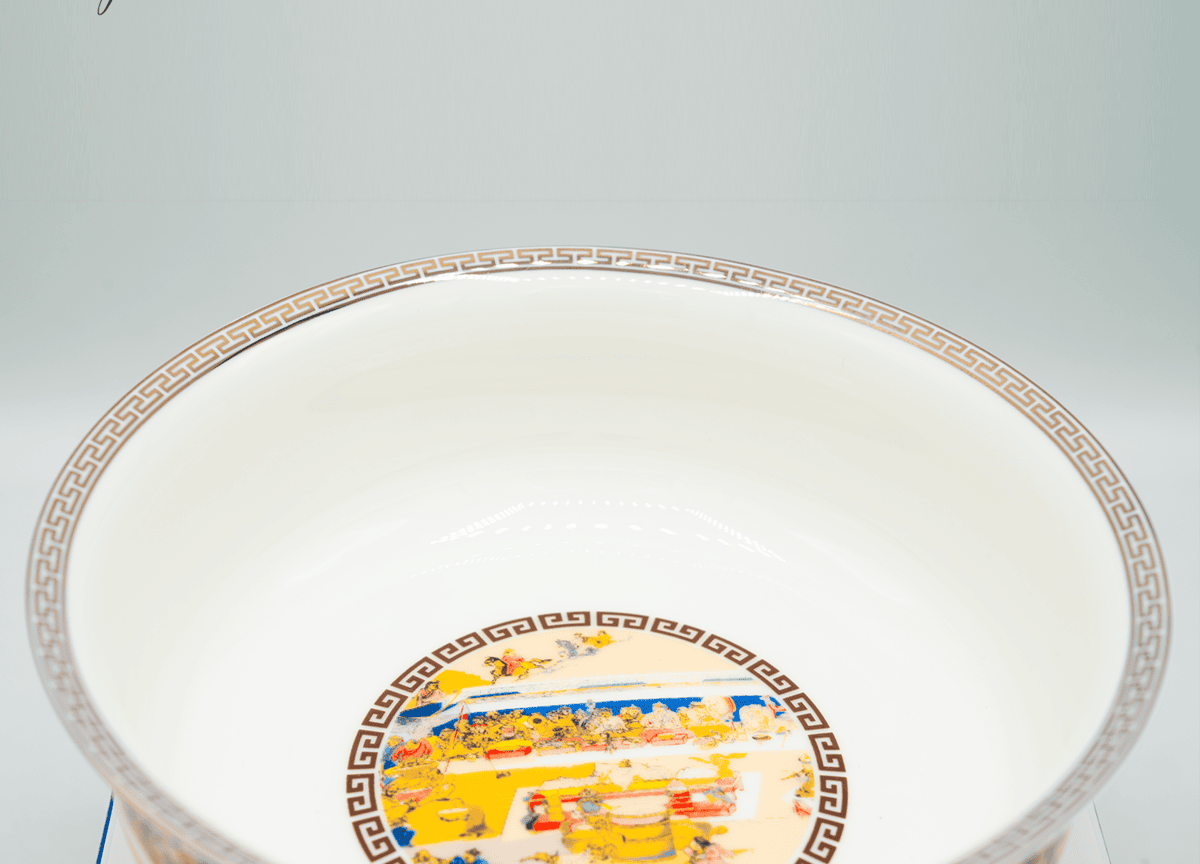 A decorative porcelain bowl with colorful illustrations on the inner bottom, featuring intricate patterns. The rim has a geometric border, and a signature is visible on the top left corner. The background is plain white.