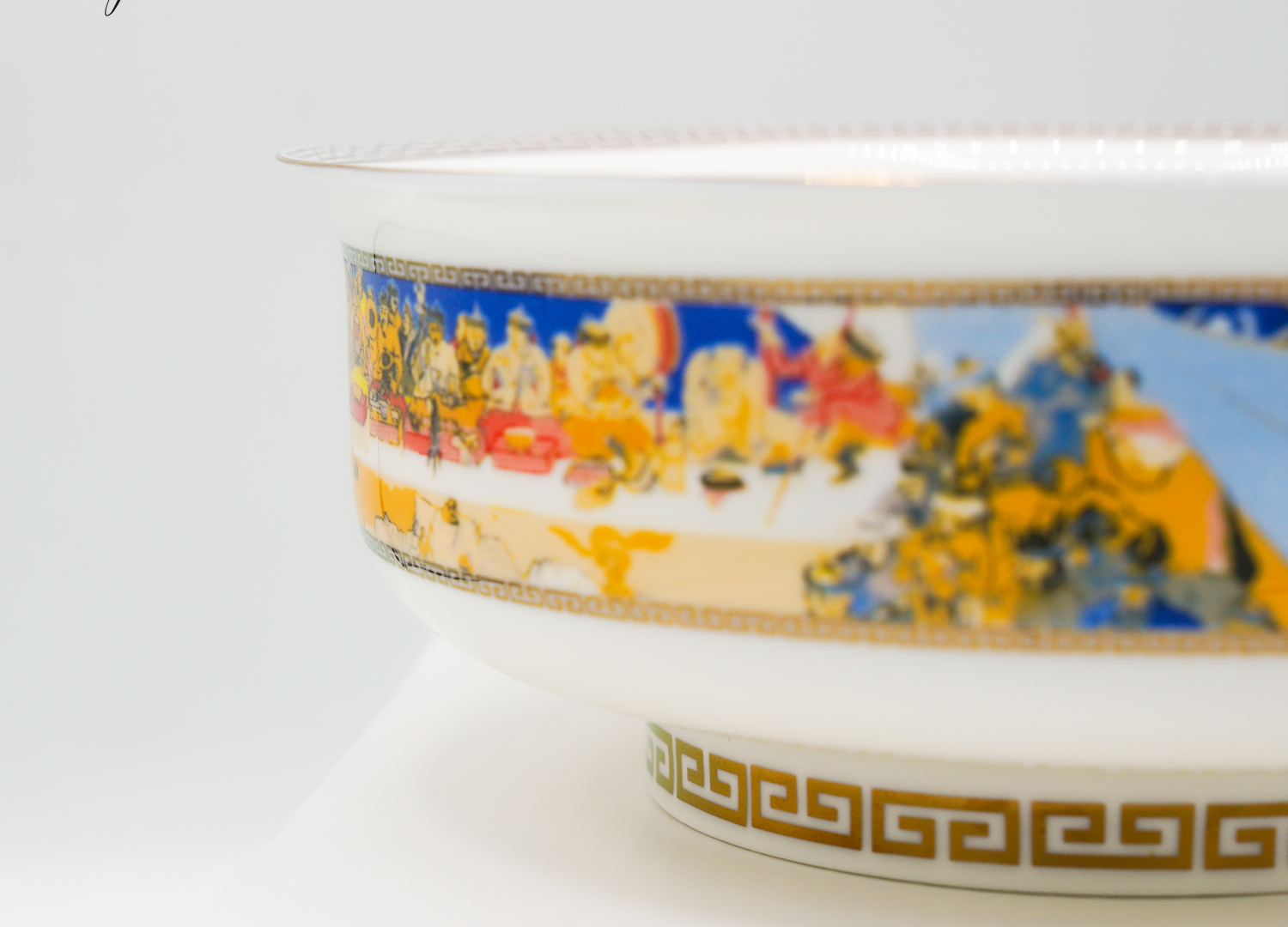 Close-up of a decorative porcelain bowl featuring intricate colorful artwork with gold accents and geometric patterns along the rim. The bowl is displayed on a white surface against a light background.