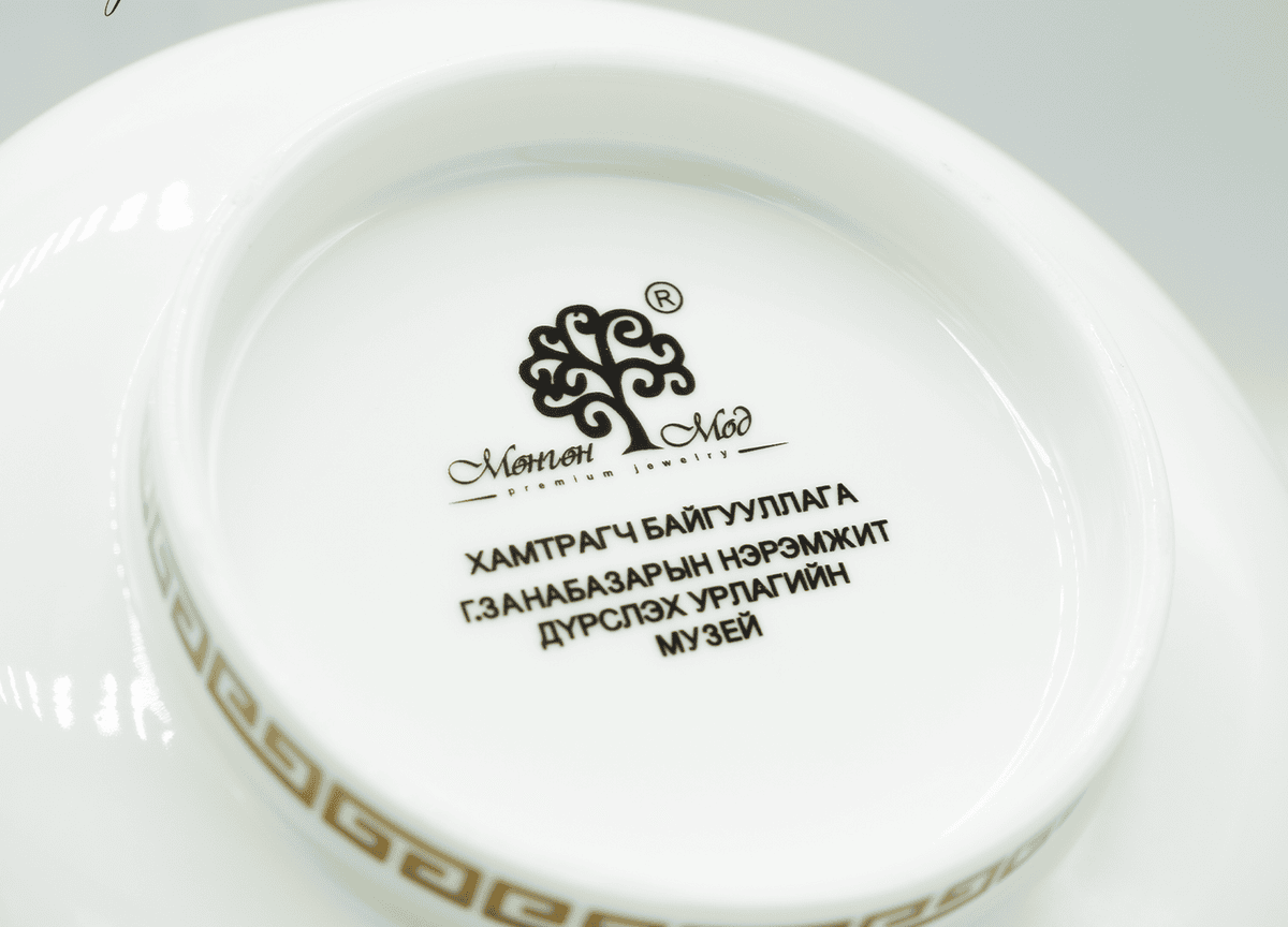 A ceramic plate with decorative border features Mongolian script and a logo in the center, set against a white background.