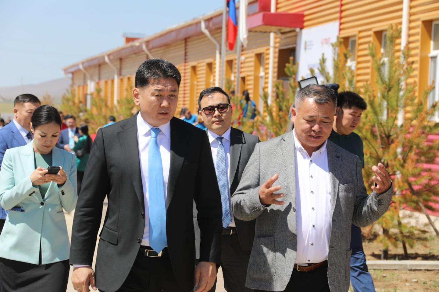 A group of people walks outside near a yellow building with small trees beside them. The man on the right gestures as he speaks, while the others listen. Another man and a woman walk slightly behind, with the woman looking at her phone.