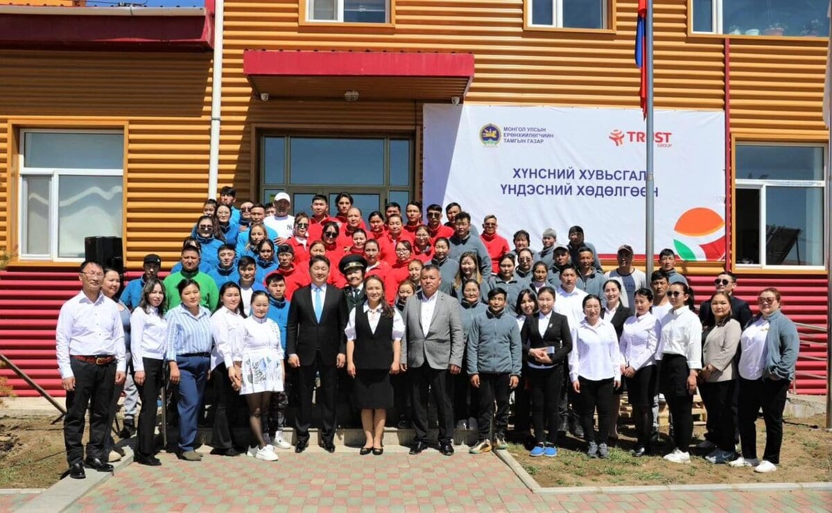 A large group of people stand in front of a building with red and yellow siding. They are smiling and diverse in attire. There is a banner on the building with text and logos. The setting appears to be outdoors on a sunny day.