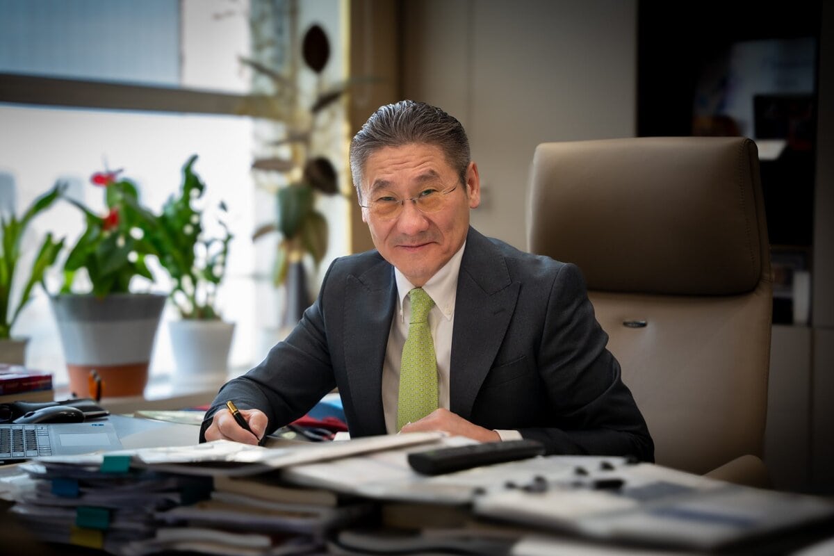 A man in a suit and tie sits at a desk covered with papers, smiling at the camera while holding a pen. There are potted plants and a large window in the background, creating a professional office setting.