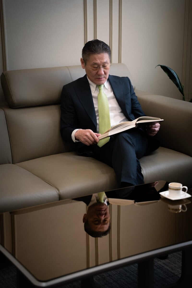A man in a suit and green tie sits on a beige sofa reading a book. His reflection appears on the glass coffee table in front of him, which holds a cup and saucer. A plant is in the background.
