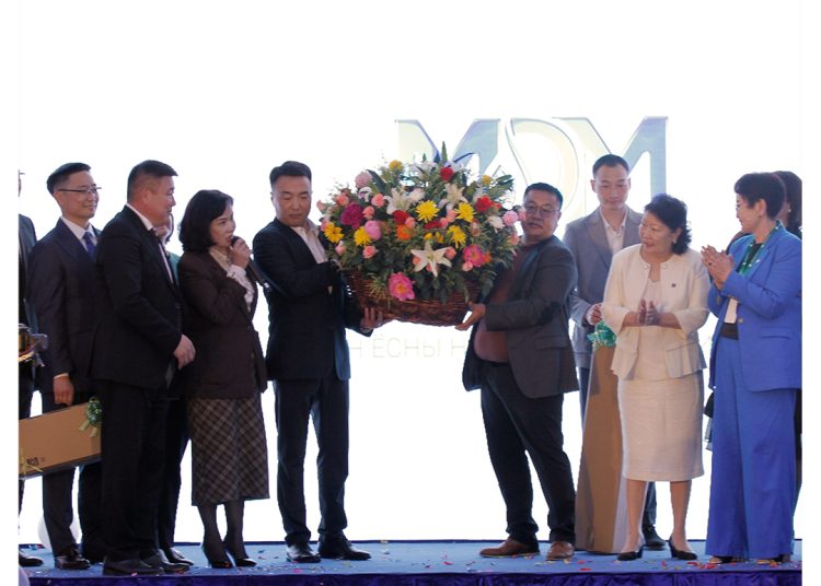 A group of people in formal attire stands on a stage. Two men hold a large basket of colorful flowers. Other people stand nearby, clapping and smiling. Balloons and confetti are on the floor. The MDM logo is visible in the background.