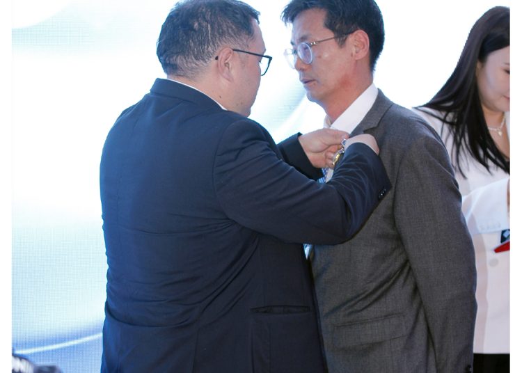 A man in a suit pins a medal or badge onto another man's suit jacket during a formal ceremony, with two women standing in the background. The MDM Ampolle Pharma logo is visible in the top right corner.