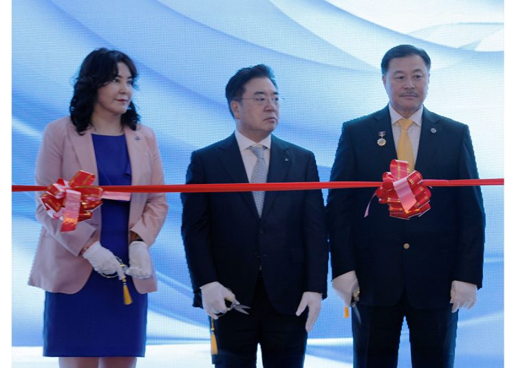 Three people in formal attire stand behind a red ribbon with bows, preparing for a ribbon-cutting ceremony. They are holding scissors and wearing white gloves, with a blue and white backdrop behind them.