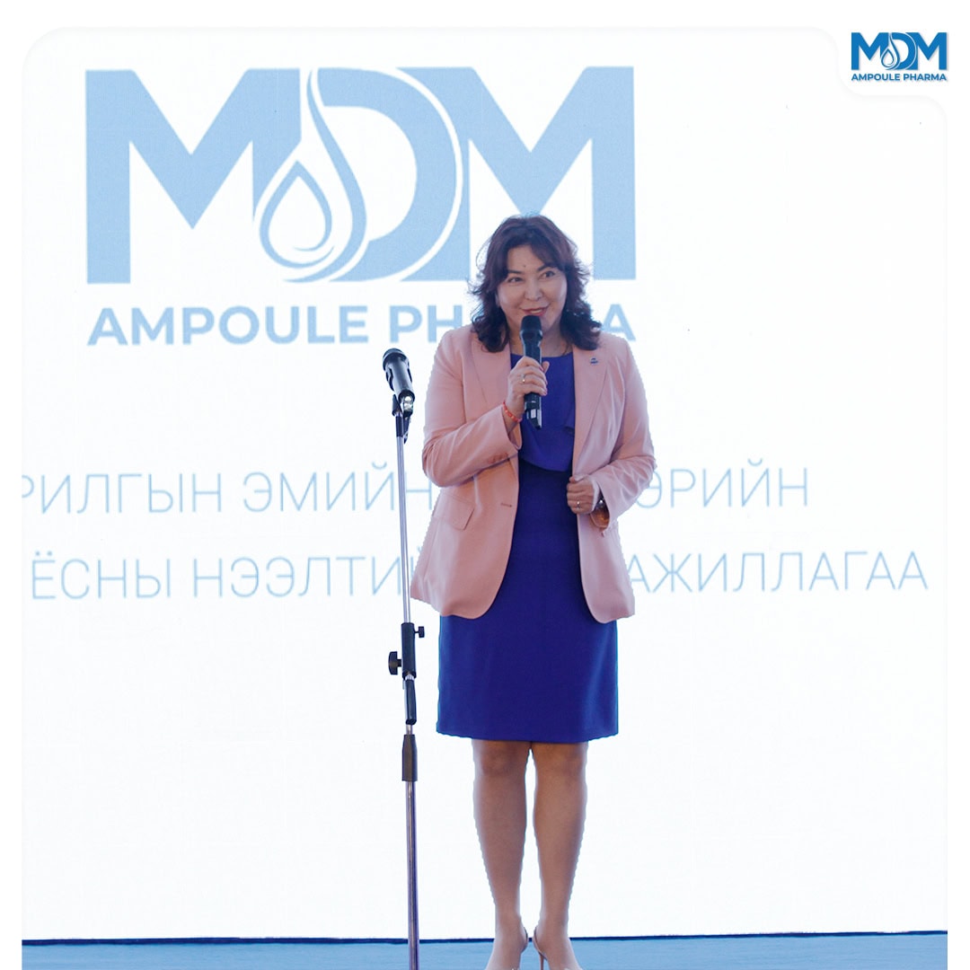 A woman in a pink blazer and blue dress speaks into a microphone on stage in front of a white background with the "MDM Ampoule Pharma" logo and text.