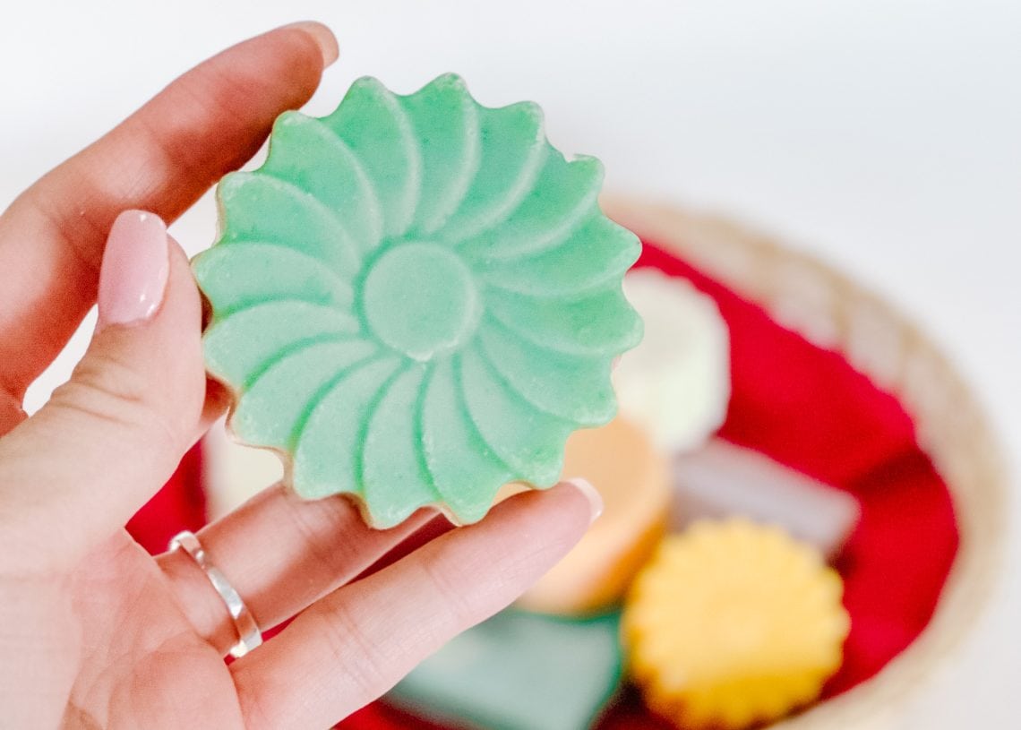 A hand with light pink nail polish holds a green, flower-shaped soap. In the background, a wicker basket with a red lining contains other colorful soaps.