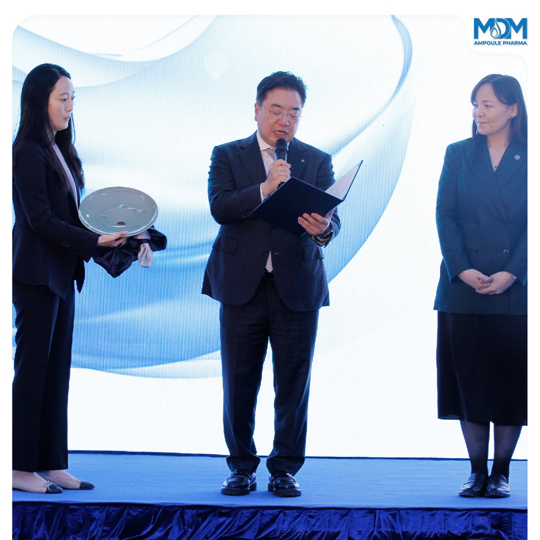 Three people in formal attire stand on a stage; the man in the middle reads from a document, while the woman on the left holds a round object, and the woman on the right stands with her hands clasped.