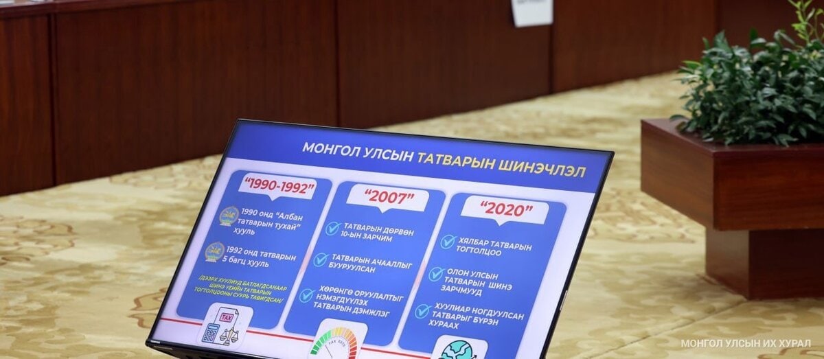 A digital display screen shows a timeline and key points about Mongolian tax reforms from 1990 to 2020, featuring colorful icons and Mongolian text in a conference room setting.