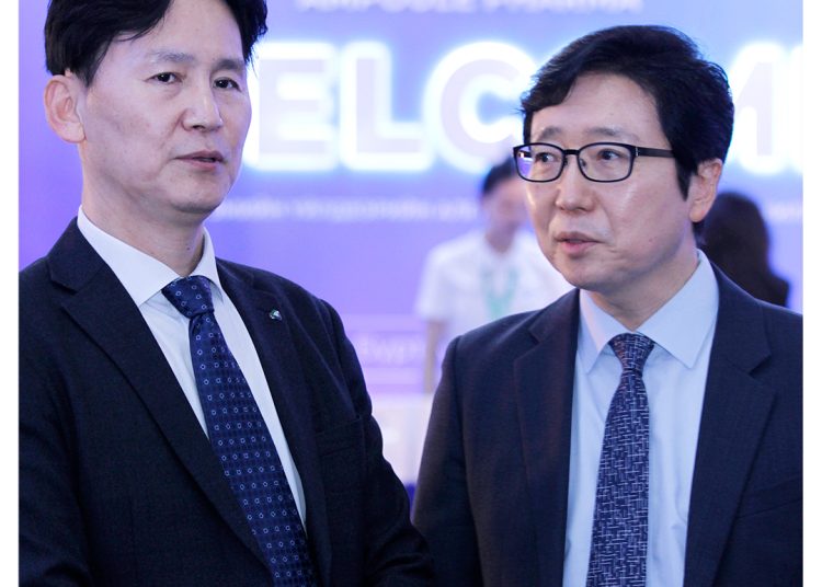 Two men in suits and ties stand together at an event with a blue "WELCOME" sign in the blurred background. The logo "MDM AMPOULE PHARMA" is visible in the top right corner.