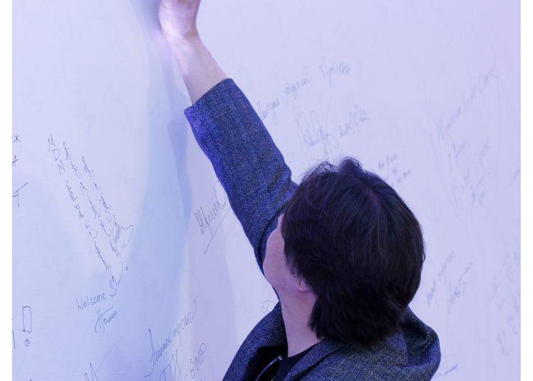 A person in a gray blazer writes a signature on a white wall covered with various handwritten signatures and messages. The logo "MDM Arnolfe Tindana" appears in the top right corner.